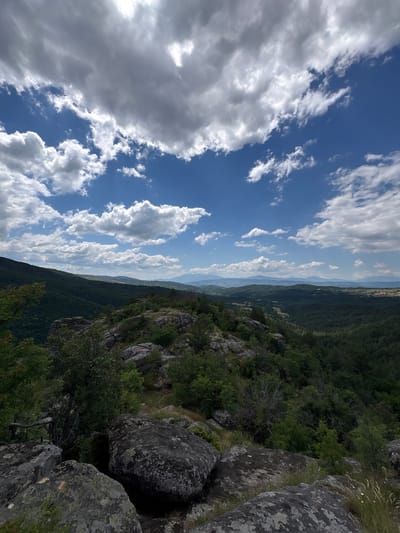 The image depicts a wide, panoramic view from a rocky, elevated vantage point overlooking a valley and distant mountains. The foreground is dominated by large, weathered grey rocks, some covered in lichen, with sparse green vegetation and grass growing between them. A wooden railing is partially visible on the left.
Beyond the immediate rocky outcrop, the landscape transitions into a dense, verdant forest that covers the slopes of rolling hills. The forest canopy is a deep green, suggesting mature trees. Further in the distance, the terrain softens into gentler, layered hills with lighter green and yellow patches, indicative of fields or sparser vegetation. The furthest horizon is marked by a range of hazy, blue mountains under a bright sky.
The sky itself is a prominent feature, with a vibrant blue expanse dotted with dramatic, white and grey cumulus clouds. The clouds are dynamic, with some appearing bright and fluffy, while others are darker and more substantial, suggesting a partly cloudy day with potential for passing showers.
The location is identified as the city of Dolno Drianovo, Bulgaria. The scene conveys a sense of natural beauty and a rugged, untamed environment. There are no people or animals visible in the image. The atmosphere is serene and majestic, with a focus on the vastness of the natural landscape.
