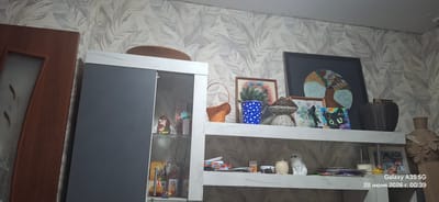 The image shows an interior scene, likely a living room or display area, featuring a wall with a leaf-patterned wallpaper. A tall, narrow cabinet with a gray sliding door is positioned on the left, partially open to reveal items on shelves within. Above the cabinet, a large, round, wooden decorative plate is placed on top. To the right of the cabinet, a white shelving unit displays various decorative objects. These include a wicker basket, a blue and white polka-dot pot with a small green plant, framed artwork, and a woven mushroom-shaped object. Further to the right, a dark woven basket and a tall, textured decorative vase are visible. The lower shelf of the white unit holds a small white cat figurine, a wooden container, and several papers. A timestamp in the bottom right corner indicates "Galaxy A35 5G 28 июня 2026 г. 00:39," suggesting the photo was taken in Belorechensky, Russia, on June 28, 2026, at 00:39. The overall impression is of a home environment with personal decorative items.