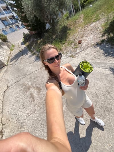 A blonde woman wearing sunglasses, a white athletic outfit, and white sneakers takes a selfie while holding a rolled-up yoga mat and a water bottle. She is standing on a paved path that slopes downwards. To her left, a white building with balconies is visible, and to her right, there is a grassy slope with trees. The scene appears to be outdoors in a residential or resort area, possibly in Ivanovići, Montenegro, given the Mediterranean-style architecture. The lighting suggests it is daytime, with strong sunlight casting shadows.