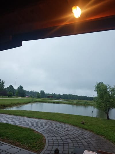 An overcast, possibly rainy day in Antāni, Latvia, is depicted. The foreground shows a paved walkway made of grey interlocking bricks, curving from the bottom left towards the right. Adjacent to the walkway is a patch of green grass. In the middle ground, a calm body of water, likely a pond or small lake, is visible, reflecting the overcast sky. Along the far edge of the water, reeds and tall grass grow, and beyond that, a line of dark green trees forms a dense forest. To the left, a red car is parked on a grassy area, and a flag pole stands nearby. A small wooden building with a dark roof is situated on the right side of the water. In the upper portion of the image, a light fixture with a yellow bulb is attached to a wooden overhang, casting a warm glow and lens flare. A single duck is standing on the grass near the edge of the water on the right side of the frame. The overall scene suggests an outdoor recreational area or a park-like setting.