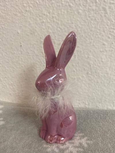 A glossy, pink ceramic rabbit figurine, adorned with a fluffy white feather boa around its neck, sits on a textured gray surface. The figurine is positioned in the foreground, facing left, with its ears upright and a subtle sheen reflecting light. The background is a textured off-white wall. The scene appears to be indoors, possibly on a shelf or table, with no specific identifiable city or location details present in the image. Sachsenheim, Germany is not depicted in this photograph.