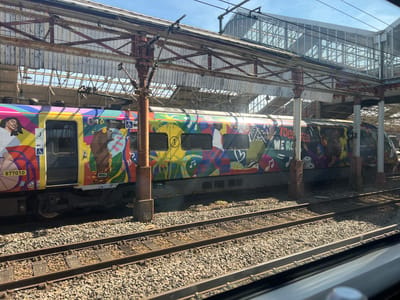 The image shows an Avanti West Coast train at Crewe railway station in Crewe, Cheshire, UK. The train has a colorful design with figures and images. Tracks run in the foreground. Above the train is the steel framework of the station roof and a footbridge. The sky is blue and sunny.