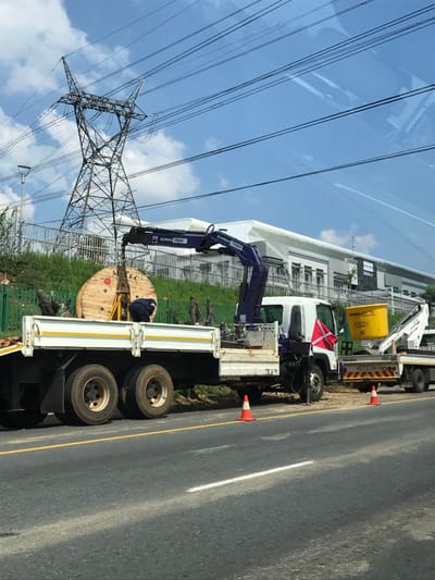 The image displays a roadside utility operation underway in Sandton, South Africa, beneath a blue sky with scattered white clouds and visible contrails. In the foreground, an asphalt road with a solid yellow line and a dashed white lane marker borders a work area. Two orange traffic cones delineate a section of the road shoulder. A white flatbed truck equipped with a blue hydraulic crane, branded "BOWMAN CRANE," is parked on the shoulder. The crane's arm is extended and engaged with a large, light-brown wooden cable drum, labeled "MEC," which is situated on the truck's flatbed. One individual, dressed in dark workwear, is observed bent over near the cable drum, appearing to interact with it or associated equipment. Directly behind this vehicle, a second white utility truck, identified as "ISUZU" on its rear, features a yellow elevated bucket or container mounted on its rear platform. In the background, a grassy embankment, topped with a metal fence, supports a prominent metal lattice power line pylon. Multiple overhead power lines span the upper portion of the scene. Beyond the embankment, a modern, light-colored building complex with white walls and a gray roof is visible.