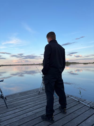 A person is standing on a wooden dock next to a body of water. The person is wearing dark clothing and shoes with yellow accents. A fishing rod rests on a tripod near the dock's edge. The sky is visible with reflections of clouds in the water. The location is outdoors, likely in or near Khryashchevka, Russia, as indicated by the prompt.