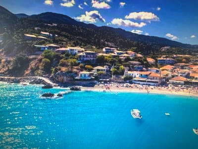 This media file captures a picturesque aerial view of a coastal village nestled at the foot of a lush, forested mountain. The scene is bathed in bright sunlight, suggesting a clear, warm day, likely midday or early afternoon, given the strong illumination and scattered white clouds against a vibrant blue sky.
The village itself is characterized by numerous buildings with terracotta-tiled roofs, clustered along the hillside and extending towards a sandy beach. These structures appear to be a mix of residential homes and possibly hotels or guesthouses, painted in light colors. The hillside is densely covered with green trees and shrubs, with some of the buildings seemingly built directly into the slope.
A busy beach is visible, dotted with numerous people enjoying the sunny weather. Some are sunbathing on the sand, while others are in the shallow water, engaged in various beach activities. The water is a striking, clear turquoise, with gentle waves lapping against the shore. A few small boats, including a white motorboat, are anchored or cruising in the bay, adding to the sense of a popular vacation destination.
The presence of rocky outcrops along the coastline and the overall Mediterranean-like appearance of the architecture and vegetation hint at a location in a region with a warm climate, possibly a European coastal area. The image evokes a feeling of leisure and relaxation associated with a summer holiday. No visible text is present in the media file.
