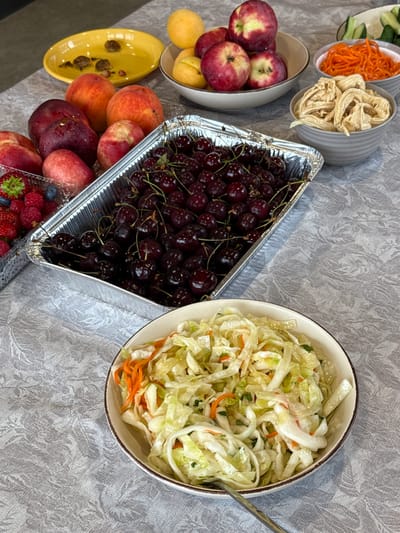 A close-up, eye-level shot shows a table covered with a grey, patterned tablecloth. Various fruits and dishes are arranged on the table. In the foreground, a white bowl with a brown rim contains a salad made of shredded cabbage and shredded carrots. A fork rests in the salad. Behind the salad bowl, a rectangular aluminum foil container is filled with dark red cherries. To the left of the cherries, in a clear plastic container, are raspberries, strawberries, and blueberries. Further back, on the left, are several peaches and dark red plums. In the upper left corner, a yellow oval dish holds what appear to be three snails. To the right of the yellow dish, a white bowl holds apricots and red and yellow apples. Behind the bowl of apples, a gray bowl contains shredded carrots, and another gray bowl holds a beige, stringy food item. In the background, a white plate holds sliced cucumbers. The setting is indoors, with a neutral background that suggests it is a dining area. The image does not provide any information about the location in the city of Bulatovo, Russia.