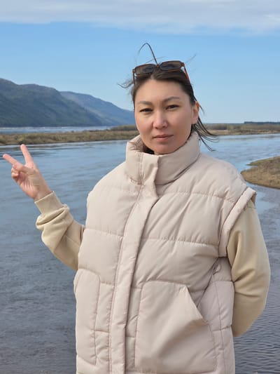 The image shows a woman standing outside, likely in Lensk, Russia, with a river and landscape in the background. The woman is wearing a light-colored quilted vest over a tan sweatshirt. Sunglasses are perched on her head. She is making a peace sign with her hand. The background consists of a wide river, a distant shoreline with vegetation, and hills under a blue sky.