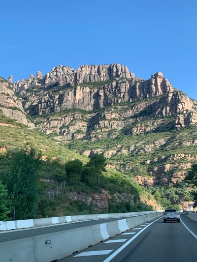 An eye-level, wide shot captures a white SUV driving on a highway with concrete barriers separating the lanes and shoulders. The highway curves to the right in the foreground, and the mountain range of Montserrat, located near Esparreguera, Spain, dominates the background. The mountain slopes are covered in green vegetation, and rocky outcrops rise sharply towards a clear blue sky. The sun is high, casting shadows on the mountain.