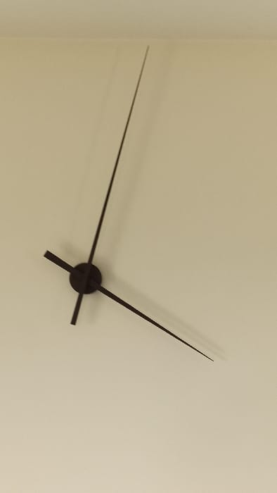 The image displays a minimalist wall clock mechanism located in Florenville, Belgium. Three black clock hands radiate from a central black circular hub against a plain, light beige or off-white wall. The shortest, thickest hand, presumed to be the hour hand, points left-horizontally. The medium-length, thinner hand, presumed to be the minute hand, points diagonally downwards to the right. The longest, thinnest hand, presumed to be the second hand, points directly upwards. Shadows are visible behind the hands on the wall, indicating a light source from the upper left. No clock face numbers, markings, people, or other objects are present in the scene.