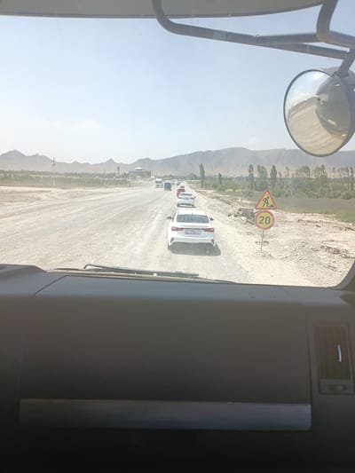 The image depicts a traffic jam on a dusty, unpaved road in a rural mountainous area. The perspective is from inside a vehicle, looking forward. Several white vans and cars are visible in a line, suggesting a queue. In the foreground, a large side mirror of the vehicle is reflected in the windshield.
Two road signs are present. One is a triangular warning sign with a symbol of a person shoveling, indicating road work or construction ahead. Adjacent to it is a circular sign with a red border and the number "20" in yellow, signifying a speed limit of 20.
The landscape features dry, light-colored earth and scattered vegetation. In the distance, rolling hills or mountains are visible under a bright, hazy sky. The lighting suggests it is daytime, likely late morning or afternoon, with the sun casting a diffused light. The presence of dust on the road and the hazy atmosphere might indicate dry weather conditions. The overall impression is one of a remote, possibly developing area where road conditions may be challenging. No people are clearly discernible in the image. There is text on the license plate of the white van in the foreground, "HR 279 AVF".