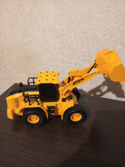 This media file shows a close-up, eye-level, side view of a yellow toy front-end loader, possibly a replica of a ZHXIMEIXIN model. The toy is positioned on a dark wooden surface, likely a table or shelf, with a textured, light-colored wall in the background. The wall has a subtle, repeating pattern and some small, reflective specks that might be glitter or dust.
The front-end loader has a black cab with tinted windows and a black engine cover. The lifting arm and bucket are bright yellow. There are visible black accents on the toy, including a safety cage around the cab, ladders on the side, and the tires. The wheels have yellow rims. The overall impression is that of a new or well-maintained toy.
There is text visible on the lifting arm of the loader. It appears to be "ZHIMEIXIN" written in black letters, with a "X" symbol also in black.
The lighting suggests an indoor setting, and the time of day is not discernible. There are no people or animals present. The scene is static, with the toy simply placed on the surface. The location context of Novotroitsk, Russia, is provided but not visually evident from the image itself.