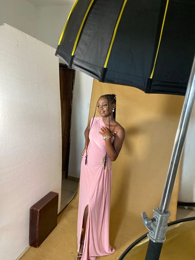 The image depicts a young woman posing for a photoshoot indoors. She is wearing a long, light pink, sleeveless dress with a high slit on the side. The dress has decorative embellishments around the waist. Her hair is styled in braids and she is wearing earrings and a watch. She is smiling and looking towards the camera, with her hands placed on her chest.
The setting appears to be a studio with a warm yellow backdrop. A large, black photographic umbrella with yellow accents is positioned to the left of the subject, suggesting professional lighting equipment is in use. A metal stand with a clamp is also visible, likely part of the lighting setup. In the foreground, a white surface, possibly a backdrop or a large styrofoam board, dominates the lower portion of the image. A dark brown, rectangular box or case rests on this white surface.
The image is captured in portrait orientation, with the subjects and scene rotated 90 degrees counter-clockwise. The lighting suggests an indoor environment, possibly during the day. The overall mood is positive, with the subject appearing confident and happy. No visible text is present in the image. The location context is Anguldi, Nigeria.