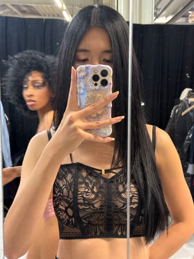 The image captures a mirror reflection of two females in an indoor setting, likely a dressing room or backstage area, in Herentals, Belgium. A female with long, straight, dark hair parted centrally is positioned in the foreground, holding a smartphone with a multi-lens camera and an iridescent, textured case in her right hand, obscuring the upper portion of her face. Her visible fingers display manicured nails with a light pink base and white tips. She wears a black lace bralette with thin straps and a small gold-colored decorative element at the chest. In the background, reflected behind the primary subject, a second female with voluminous, dark, curly hair is partially visible. She appears to be looking towards the left with a slightly open mouth, exhibiting light-colored eye makeup. The environment includes dark curtains and a clothing rack with garments visible in the reflection. An overhead light source is also reflected at the top center.