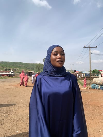 A woman wearing a blue hijab and dress stands outside in Naraguta Hausawa, Nigeria. She is the primary subject. In the background, other people wearing long garments are walking along a dirt road. Buildings are visible in the distance, along with a truck. Power lines are overhead. The sky is cloudy.