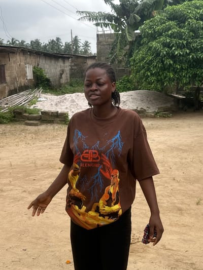 A young Black female stands in an outdoor, dusty environment in Ganyingbo, Nigeria. She is wearing a brown t-shirt with a graphic design featuring lightning bolts and figures, and black pants. Her right hand is slightly extended with open fingers, while her left hand holds a red and blue checkered cloth. In the background, there are low-rise buildings constructed with concrete blocks, some with corrugated metal roofs. Vegetation, including palm trees and leafy trees, is visible, along with piles of sand and building materials. The ground is a mixture of sand and dirt. The sky is overcast.