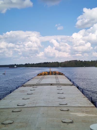 The image depicts a view from the deck of a large barge or ship traveling on a body of water, with a forest visible in the distance. The deck of the ship is large and gray, with a series of round hatches or access points. The water is a deep blue color, reflecting the sky above, which is a bright blue with puffy white clouds. Another ship or barge is visible further down the waterway, and navigation markers are also visible. The location is Kurikaevo, Russia.