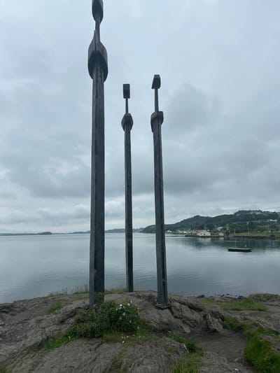 The image shows the "Swords in the Rock" monument in Stavanger, Norway. Three large, dark-colored sword sculptures are planted in the rocky ground on a hill overlooking a body of water. The largest sword is in the foreground and to the left, with two smaller swords to its right. The swords have a rough, metallic texture and sharp edges. The water is calm and reflects the overcast sky. In the background, there are green hills with scattered houses and trees, and a small dock with a yellow buoy is visible on the water. The sky is cloudy and gray.