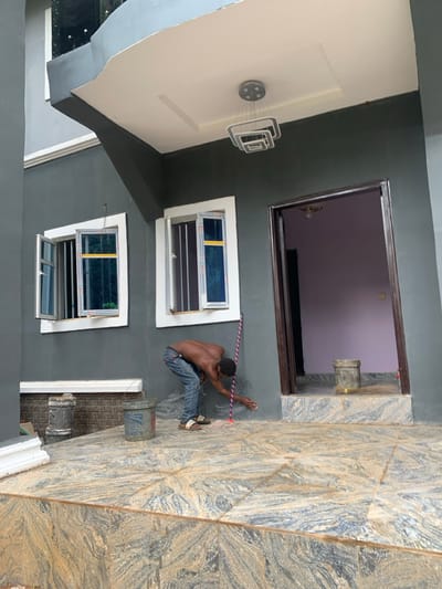 This image captures an exterior scene at a building in Amaeshi, Nigeria, depicting a single male individual engaged in maintenance or finishing work. The subject, a dark-skinned male wearing blue jeans and slide sandals, is shirtless and bent at the waist, facing the dark grey wall of the building. His right hand is near the base of the wall, possibly cleaning or applying a substance. A red and white striped mop is leaning against the wall beside him.
The building features a dark grey painted facade with white window frames. Two ground-level windows are visible, both partially open, revealing blue-tinted or covered glass and internal security bars. To the right of the subject, an open doorway with a dark brown frame leads into a room with light purple walls and a ceiling light fixture. The outdoor area is paved with large, polished rectangular tiles displaying a variegated pattern of grey, beige, and brown. A step, also tiled, leads up to the doorway. Two plastic buckets, one grey and one blue, are situated on the tiled floor near the subject. Above, a white ceiling overhangs the patio, from which a modern, multi-tiered rectangular chandelier with chrome accents is suspended. A broom is visible on the tiled floor just inside the open doorway.