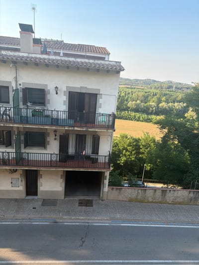 The image captures a view from an elevated position looking down onto a three-story building in Hostalric, Spain. The building's exterior is primarily white with stone accents around the windows and balconies. It features multiple balconies with black metal railings, some with chairs and laundry visible. The top of the building is adorned with a red tiled roof and a chimney.
Beyond the building, a lush green landscape unfolds. A field with agricultural patterns is visible, surrounded by dense trees and rolling hills that recede into the distance under a clear blue sky. A road runs in front of the building, with a sidewalk and a low concrete wall separating it from the property. A light-colored car is parked beyond the wall, partially obscured by trees. The scene suggests a residential area integrated with the natural environment.