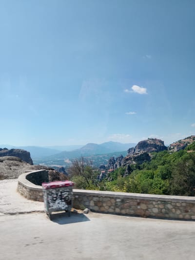 The image depicts a panoramic view of the Meteora rock formations near Kalambaka, Greece. The sky is a clear, bright blue with a few wispy white clouds. In the foreground, a winding road curves to the left, bordered by a stone wall constructed of irregular, rounded stones. To the left of the road, a trash can is visible, covered in graffiti. Beyond the wall, the landscape opens up to reveal the dramatic, towering rock pillars characteristic of Meteora, some topped with monasteries. The rocks are covered in lush green trees and vegetation, creating a stark contrast between the natural formations and the built structures. In the distance, rolling hills and mountains fade into a hazy blue, suggesting a vast and expansive environment. The overall impression is one of natural beauty and architectural marvel.