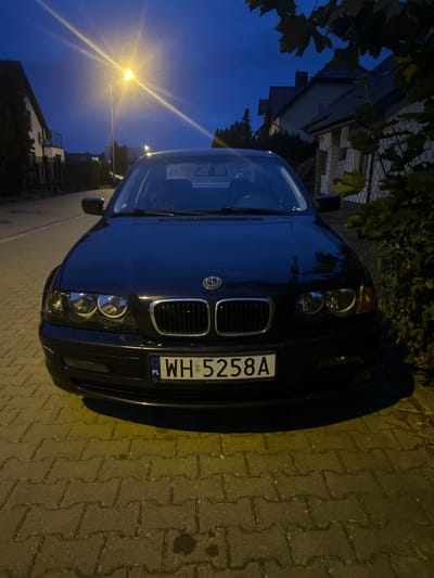 A black BMW sedan is parked on a cobblestone sidewalk at dusk, facing the camera. The car's headlights are on, illuminating the street. The license plate reads "WH 5258A" and has a Polish flag emblem. In the background, there are houses with pitched roofs and a street lamp that casts a bright light. Trees and bushes are visible on the right side of the image. The sky is dark blue, indicating it is either early morning or late evening. The scene appears to be in a residential area in the city of Rumia, Poland.