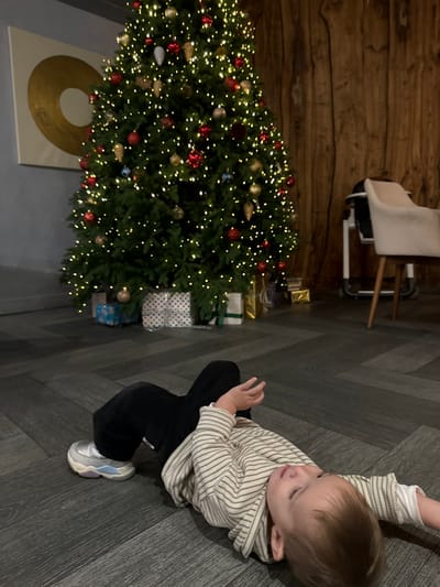 An interior scene in Kalinovo, Russia, shows a young child lying supine on a dark, herringbone-patterned floor. The child wears a light-colored striped long-sleeve shirt, dark corduroy pants, and a single light-colored sneaker on their right foot. Their head is turned slightly to the left, looking upwards, with the right arm bent and hand near their chest, and the left arm extended. In the background, a large, illuminated Christmas tree stands prominently, decorated with numerous warm white lights, red, gold, and blue ornaments. Several wrapped gifts are arranged at the base of the tree. To the left behind the tree, a white canvas with a large gold circular design is mounted on a light gray wall. To the right, a vertical wooden paneled wall is visible, along with a cream-colored upholstered chair and a white baby high chair on wheels. The lighting is dim, primarily emanating from the Christmas tree.