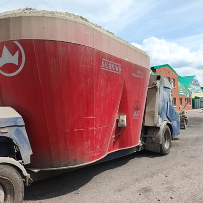 This media file shows a red Siloking SelfLine Premium 2215 feed mixer wagon, partially filled with light-colored material, possibly feed or silage. The wagon is attached to a blue vehicle, suggesting it is self-propelled. The scene appears to be an outdoor agricultural or industrial setting, possibly a farm or feed processing facility, with a gravel or dirt ground surface. In the background, there are brick buildings with green roofs, and a clear sky with scattered white clouds, indicating daytime weather. Visible text includes "SILOKING", "SelfLine", "Premium 2215", and a "4.0" logo on the side of the red wagon. There are no people visible in the image. The overall impression is a functional piece of agricultural machinery in use or awaiting use on a sunny day.