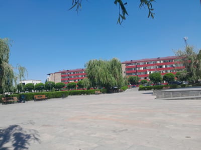 An aerial perspective of a public square in Artashat, Armenia, shows a vast, paved area with a few benches and a planter box. In the background, a multi-story red building with white-trimmed windows lines the square. Trees and shrubs provide a natural border, and a cell tower stands tall against the clear blue sky. The scene is bathed in bright sunlight, casting shadows across the pavement.