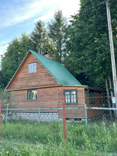 A two-story wooden house with a green metal roof is visible in the image, situated in a rural or suburban setting. The house features several windows with green trim, a brick chimney, and is surrounded by lush green trees and tall grass. A chain-link fence runs in front of the property, and a concrete utility pole with electrical wires is visible on the right side of the frame. The sky is partly cloudy with a light blue hue. The house appears to be in good condition, with the wooden exterior showing signs of age and weathering. The overall scene depicts a serene, natural environment.