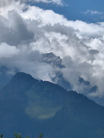 The image depicts a majestic mountainous landscape. In the foreground, a dark blue mountain slope is visible, covered with a dense forest of coniferous trees. A patch of light green meadow can be seen on the upper part of this slope. Further in the background, jagged, snow-capped peaks emerge from a layer of dramatic, white and grey clouds. The sky above is a vibrant blue, with scattered white clouds. The overall impression is one of natural grandeur and rugged beauty. The location is described as Siumsa, India, implying this is a view within or near that geographical area. No people or specific actions are discernable.
