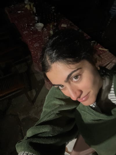 A close-up, high-angle selfie is taken by a young woman with dark hair and eyes. She is wearing a green, textured jacket over a striped shirt. Her head is tilted, and she is looking directly at the camera with a subtle smile. The background is dimly lit, showing a table covered with a red tablecloth. On the table, there are several bottles, a white bowl, and possibly some discarded cigarette butts. The overall environment appears to be outdoors or in a poorly lit indoor space. The location is identified as Felgueiras, Portugal.