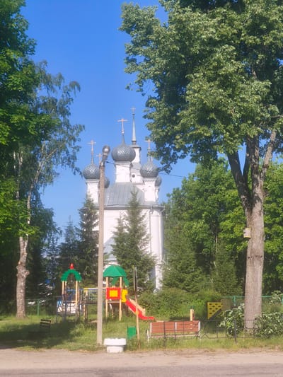The image depicts a bright, sunny day in the city of Kady, Russia. In the center of the frame, a white church with multiple grey domes topped with golden crosses stands prominently. In front of the church, a colorful children's playground is visible, complete with slides, swings, and climbing structures. To the right of the playground, a large tree with dense green foliage dominates the scene, its trunk bearing a small yellow birdhouse. The ground is covered in lush green grass and appears to be a public park or recreational area. The sky is a clear, vibrant blue.