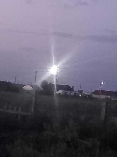 The image depicts a rural landscape at dusk or dawn, likely in Қозыкеткен ауылдық округі, Kazakhstan. A bright, starburst-like light source dominates the upper right portion of the sky, suggesting a streetlamp or a strong artificial light. In the middle ground, several low-rise buildings are visible, including one with a dark roof and a chimney, another with a red roof, and a structure covered by a white tarp. The foreground is filled with dark vegetation, possibly grass and bushes, and a wooden fence or railing. Several utility poles are scattered throughout the scene. The sky is a soft, hazy purple with some faint cloud formations. The overall impression is one of a quiet, rural setting illuminated by artificial light during a low-light condition.