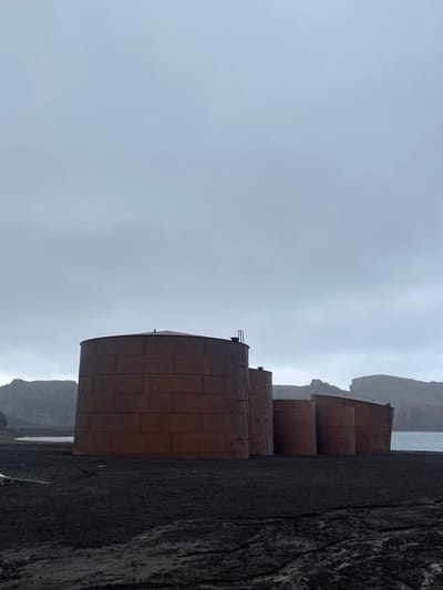 Four large, cylindrical, rusty-brown storage tanks stand in a row along a dark, gravelly terrain. The sky is overcast and gray, blending with distant, hazy hills and a body of water. The tanks appear to be old and weathered, positioned close to the edge of the land, giving a sense of isolation and industrial presence.