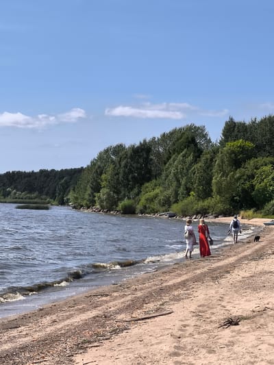 In the city of Ostrov Im. Belova, Russia, on a sunny day with scattered clouds, three individuals and a dog are walking along a sandy beach adjacent to a body of water. The water, appearing dark and somewhat choppy, meets the shore with small waves. A dense, green forest lines the opposite side of the water, with trees extending to the horizon. The beach itself is composed of sand mixed with small pebbles and driftwood.
The first individual, from left to right, is wearing a white dress and a straw hat, carrying a white backpack. The second individual is wearing a long, red dress and carrying a tan bag. The third individual, a male, is wearing a light-colored shirt and shorts, a backpack, and is holding a leash attached to a small, black dog walking beside him. The three figures are walking away from the viewer towards the right side of the frame. In the distance, several small, dark boats are pulled up on the shoreline.