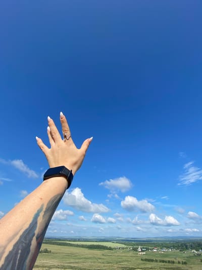 An arm with a tattoo and a smart watch extends upwards towards a clear blue sky in Andreevka, Russia. The hand is open, fingers spread, with painted fingernails and a wave-shaped ring on the middle finger. In the distance, a green landscape with scattered buildings, trees, and a church can be seen under a bright sky with scattered white clouds.