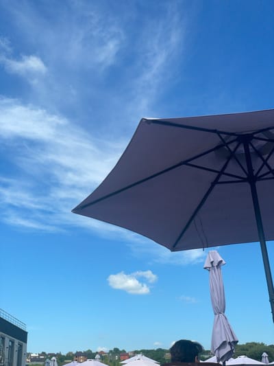 In the city of Oleshyn, Ukraine, an outdoor scene unfolds beneath a bright blue sky dotted with wispy white clouds. In the foreground, a large, dark umbrella is angled upwards, its frame clearly visible. To the right, another umbrella is closed and covered, standing upright. A person's head and shoulders are visible in the lower right corner, facing away from the camera. In the background, a building with a railing can be seen on the left, and beyond it, a line of houses and trees stretches across a gentle hill. Several other closed umbrellas are scattered throughout the scene.