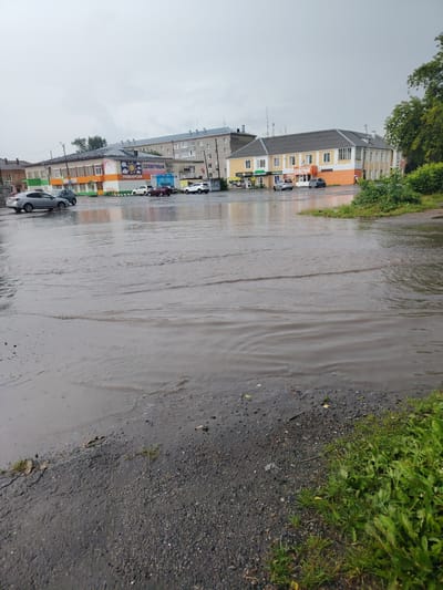 The image depicts a flooded street in Bulanaš, Russia. The scene shows a wide expanse of water covering the asphalt, with ripples indicating it is a fluid body. Several cars are parked or driving through the water. On the left side of the flooded area, there are buildings with signs, one of which reads "СОЛНЕЧНЫЙ" (Solnechny) and "УНИВЕРСАМ" (Universam). Another building on the right has a sign that appears to say "БУМЕРАНГ" (Bumerang). The weather appears overcast and possibly rainy, contributing to the flooding. Green vegetation is visible on the right side of the image, bordering the flooded street. The overall impression is one of a town experiencing significant rainfall and the resulting water accumulation on its streets. The time of day is not explicitly clear due to the overcast sky, but it appears to be daytime. There are no visible people in the image, only the presence of vehicles and buildings.