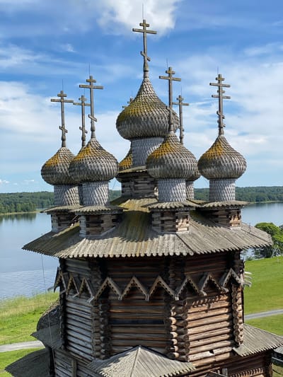 An eye-level, close-up, vertical shot captures the upper portion of the Church of the Transfiguration, a wooden Orthodox church located on Kizhi Island in the Republic of Karelia, Russia. The church is constructed from logs and features multiple onion-shaped domes topped with crosses. The domes are shingled with wooden tiles, some of which appear aged and covered in moss. Several wooden crosses, some multi-barred, are prominently displayed atop the domes and other parts of the structure.
The church's wooden construction is visible in detail, with horizontal log walls adorned with decorative wooden elements, including triangular motifs. The roofs are also shingled with wooden tiles, creating a textured appearance. In the background, a body of water, likely a lake or river, stretches across the horizon, bordered by a dense, green forest. The sky above is a vibrant blue with scattered white clouds. The overall scene is bathed in natural daylight, indicating it was taken during the daytime. No people are visible in the image.