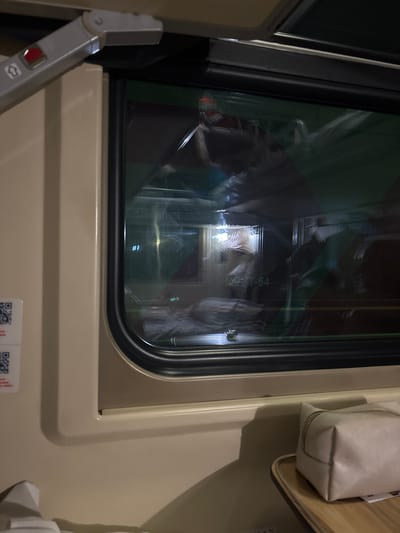 This image captures the interior of a train car at night, specifically located in the city of Tayshet, Russia.
The primary subject is a rounded rectangular train window with a black frame, viewed from the inside of the compartment. The interior of the train features light beige/off-white wall paneling. In the upper left quadrant, a gray hinged mechanism with a visible red button and an upward-pointing arrow symbol is affixed to the wall. To the immediate left of the window, a white label displaying two distinct QR codes and some red text is visible. In the lower right of the frame, a light beige zippered pouch rests on a light-colored wooden pull-down table or tray.
The window glass reflects elements from the opposite side of the train car's interior. These reflections include: a blurred individual wearing a garment with red and white stripes in the upper section; the alphanumeric text "M3T-54" near the center-bottom; and a heavily blurred figure appearing to be sleeping under a light-colored blanket or sheet on a lower bunk or seat. Numerous diffuse light sources from the interior are also reflected. A small metallic object, possibly a cup, is visible on a ledge beneath the reflected bunk. The environment outside the train is indiscernible due to darkness and strong internal reflections, indicating nighttime conditions. No direct actions or interactions are depicted.