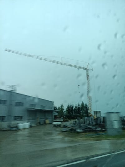 This media file shows a construction site on a cloudy day. The primary subjects are a large tower crane and a long, low industrial building. The crane, with its yellow lattice structure, dominates the upper portion of the frame, extending horizontally to the left. A hook hangs from the jib. The building, with a grey facade and several rectangular windows, is situated in the lower left. In front of the building, there are some white, possibly industrial bags, and a small truck. Beyond the building and to the right, a cluster of dark green coniferous trees can be seen. Further to the right, there are piles of construction materials, including what appear to be concrete pipes or columns. The overall scene is slightly blurred, suggesting movement, perhaps from a vehicle, and the presence of raindrops on the viewing surface, indicating it is raining. The atmosphere is muted and grey, consistent with overcast weather. No people are clearly visible, but the presence of a truck and construction materials implies ongoing activity at the site, likely related to construction or industrial operations. The location context is Šternov, Czechia, but this specific information cannot be visually confirmed from the image itself. There is no visible text in the image.