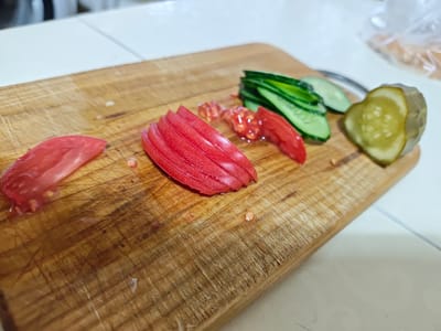 A close-up, eye-level shot shows sliced tomatoes, cucumbers, and a pickle on a wooden cutting board. The cutting board is well-worn, with visible knife marks. There are several slices of red tomato on the left, followed by some of the tomato's inner pulp and seeds. To the right of the tomatoes are several thin slices of green cucumber, and further to the right is a thick slice of pickled cucumber. The background is blurred, but appears to be a white surface, possibly a countertop or table, with a hint of plastic packaging in the upper right corner. The image was taken in Ароматненское сельское поселение, Ukraine.