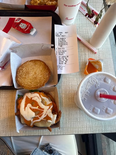 The image shows a Chick-fil-A meal on a woven surface, likely a table. The meal includes an open box with the top and bottom halves of a sandwich separated, suggesting it is being disassembled. The sandwich appears to have fried chicken, cheese, lettuce, and drizzled sauces (likely buffalo and ranch). There's a side of waffle fries and a variety of sauces including CFA sauce, buffalo sauce, ranch sauce and a ketchup packet. A Chick-fil-A branded drink cup with a red straw is also visible along with a receipt with the name Laura and the order number 4921. The receipt lists items purchased including 8 nuggets, CFA sandwiches, Spicy Chicken sandwiches, medium fries, different sodas and tea, with the order date and time of 5:06 PM. Based on the receipt mentioning "Southlake, TX" the location is most likely Chick-fil-A in Southlake, Texas, United States.