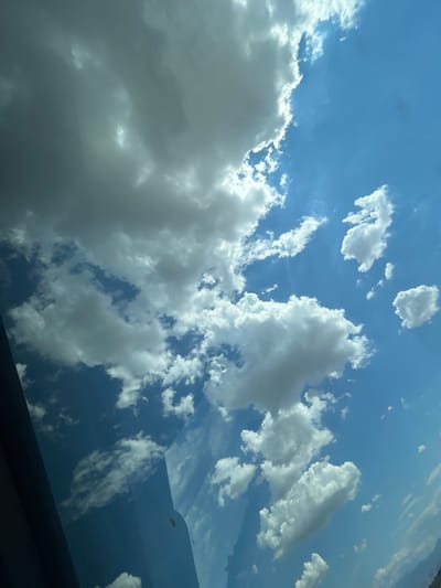 The image captures a dynamic sky with a mix of cumulus and darker, more overcast clouds against a vibrant blue sky. The perspective suggests the photo was taken from a slightly elevated position, possibly looking upwards from a vehicle or building. A dark, angled shadow or object occupies the lower left quadrant of the frame, obscuring a portion of the view. The lighting indicates it is daytime, with sunlight breaking through and illuminating some of the clouds, creating a sense of depth and volume. The scene appears to be an outdoor environment, characteristic of a region like Shtarkovo, Bulgaria, where such cloud formations and sky conditions are common. There are no people or discernible human activities depicted in the image; the focus is solely on the natural atmospheric elements. The overall impression is one of natural beauty and atmospheric change.