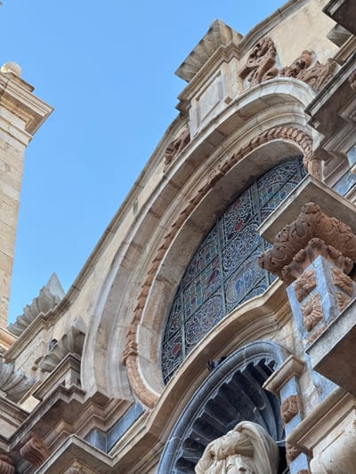The image shows a low-angle view of the architectural details of a building, likely a church, in Vinaròs, Spain. The sky is light blue, visible between the structures. The building is constructed of tan-colored stone. Ornate carvings and moldings are prominent. A section of stained glass is visible at the top. A statue or architectural detail with a draped element is also present. The architecture exhibits Gothic or Renaissance influences.