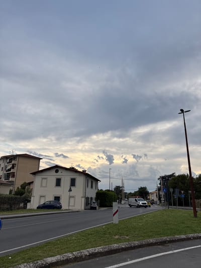 The image shows a street scene in Pisa, Italy. A two-lane road with a grassy median is bordered by buildings on one side. A two-story light-colored building with green shutters is prominent, and a blue car is parked in front of it. The sky is predominantly cloudy with streaks of light breaking through. A street lamp with a unique propeller-like design is visible. There are also traffic signs, a crossing guard, and a van parked along the road further in the distance.