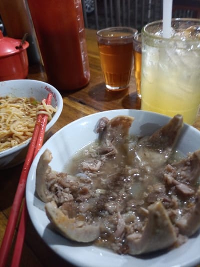 The scene depicts a meal laid out on a dark wooden table in an establishment within Ciomas, Indonesia. In the foreground, a white bowl contains a dish composed of several large, irregular meat pieces submerged in a clear, light brown broth, appearing to be a type of Indonesian bakso or stew. To the left, a second white bowl holds yellow noodles, with a pair of red chopsticks resting across its rim.

Further back on the table, several beverages are present. A large glass on the right contains a light yellow, iced drink with a white straw. To its left, two smaller glasses hold a clear, brown liquid, consistent with tea. A tall, opaque red bottle, likely containing chili sauce, stands behind the noodle bowl. Next to this bottle, a small red lidded container is also visible. The lighting is warm and appears to be artificial, suggesting an indoor setting. No individuals are visible in the frame.