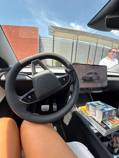 The image provides an interior view of a Tesla vehicle, with the steering wheel and dashboard prominently featured. Through the windshield, a red brick building and a modern architectural structure with slatted roofs are visible against a bright blue sky with scattered clouds.
In the background, a man wearing sunglasses and a white polo shirt is seen smiling, standing near the car. Another person, a woman with red hair, is partially visible behind the man.
Inside the car, on the center console, there are several food items including boxes of Stracciatella protein yogurt and a box labeled "Manhattan Salad."
The overall scene appears to be a sunny day, likely in an urban or commercial area, as indicated by the architectural elements. The presence of food items suggests a break or a stop during a journey. There is no indication of Les Arcs, France in the image.