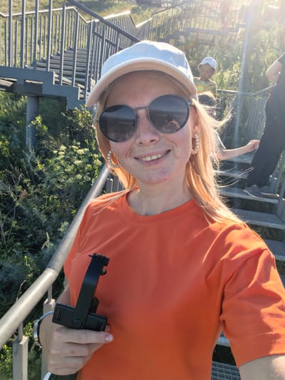 A woman wearing an orange t-shirt, a white baseball cap, and round sunglasses smiles at the camera. She holds a black handheld gimbal in her right hand. She is standing on a metal staircase outdoors, with greenery and other people in the background. The sun is shining brightly, creating lens flares in the image. The location is Akhmerovo, Russia.