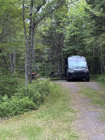 A dark-colored camper van is parked on a gravel road in a wooded area. The van is facing forward, with its windshield and front grille visible. It appears to be a modern model with tinted windows. To the left of the van, a bicycle is mounted on a rack. A metal frame structure, possibly a canopy or awning, is set up behind the van. In the background, dense green foliage and tall trees create a natural, secluded setting. The ground is covered in gravel and grass, suggesting a campsite or a rural road. The scene is likely outdoors, with natural lighting, suggesting daytime. There is no visible text in the image. The overall impression is one of a peaceful, natural environment.